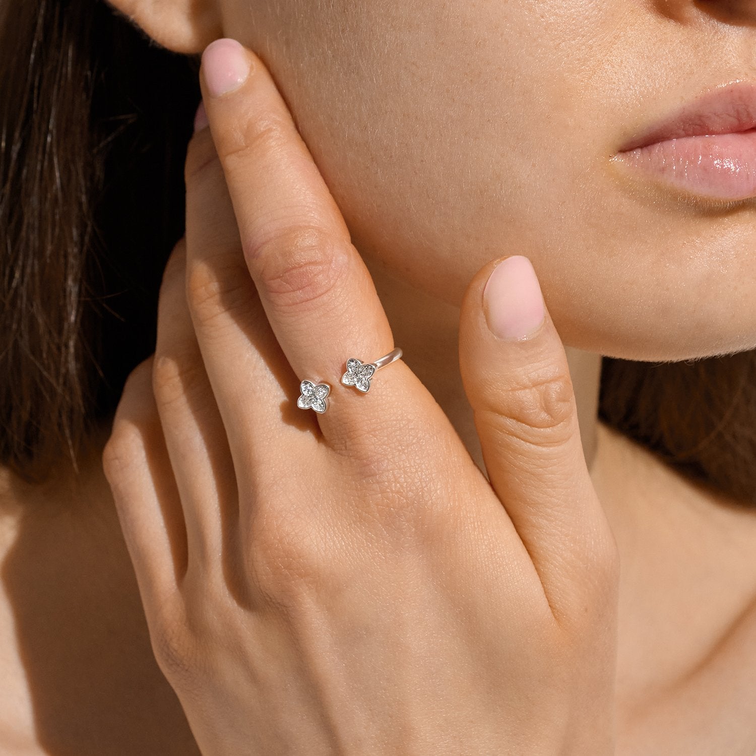 A close-up lifestyle shot of a model's hand wearing a delicate silver ring with two small, clear flower-shaped stones. The model's face and hand are illuminated by warm, direct sunlight against a dark background.