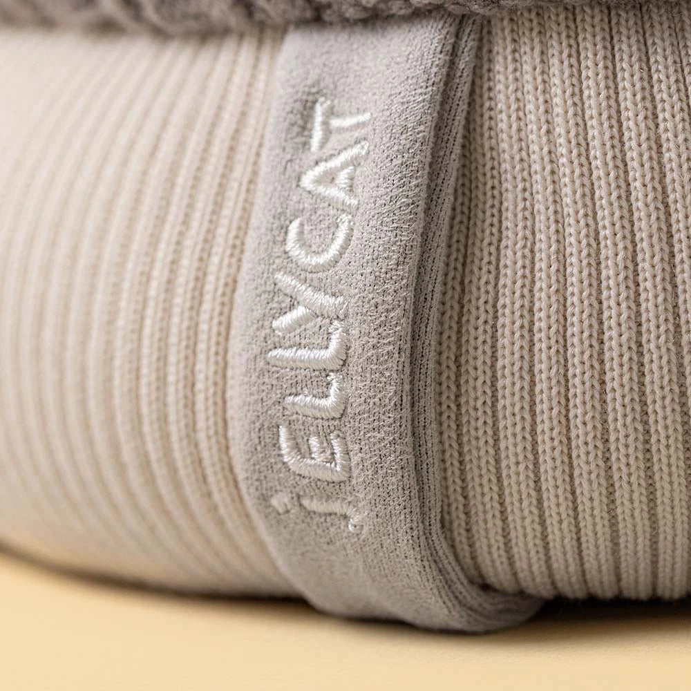 Close-up of a Jellycat amuseables pomilly hat with a gray cable-knit top, a cream ribbed bottom, a gray pom-pom, and small fabric hands and feet, shown on a white background with brand name 'JELLYCAT' embroidered vertically on a fabric tab.