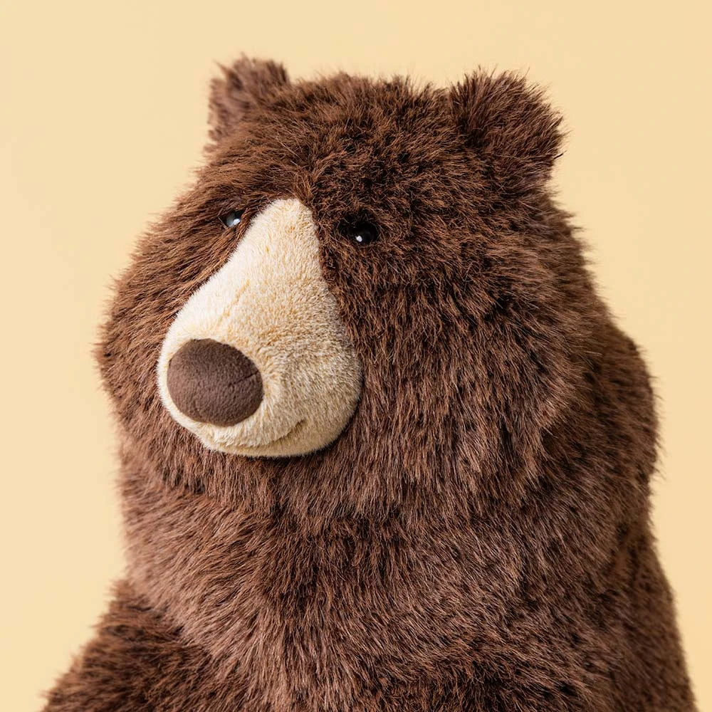 A close-up of jellycats Bizzlestomp brown plush bear toy with a cream-colored snout and dark eyes, set against a solid pale yellow background.