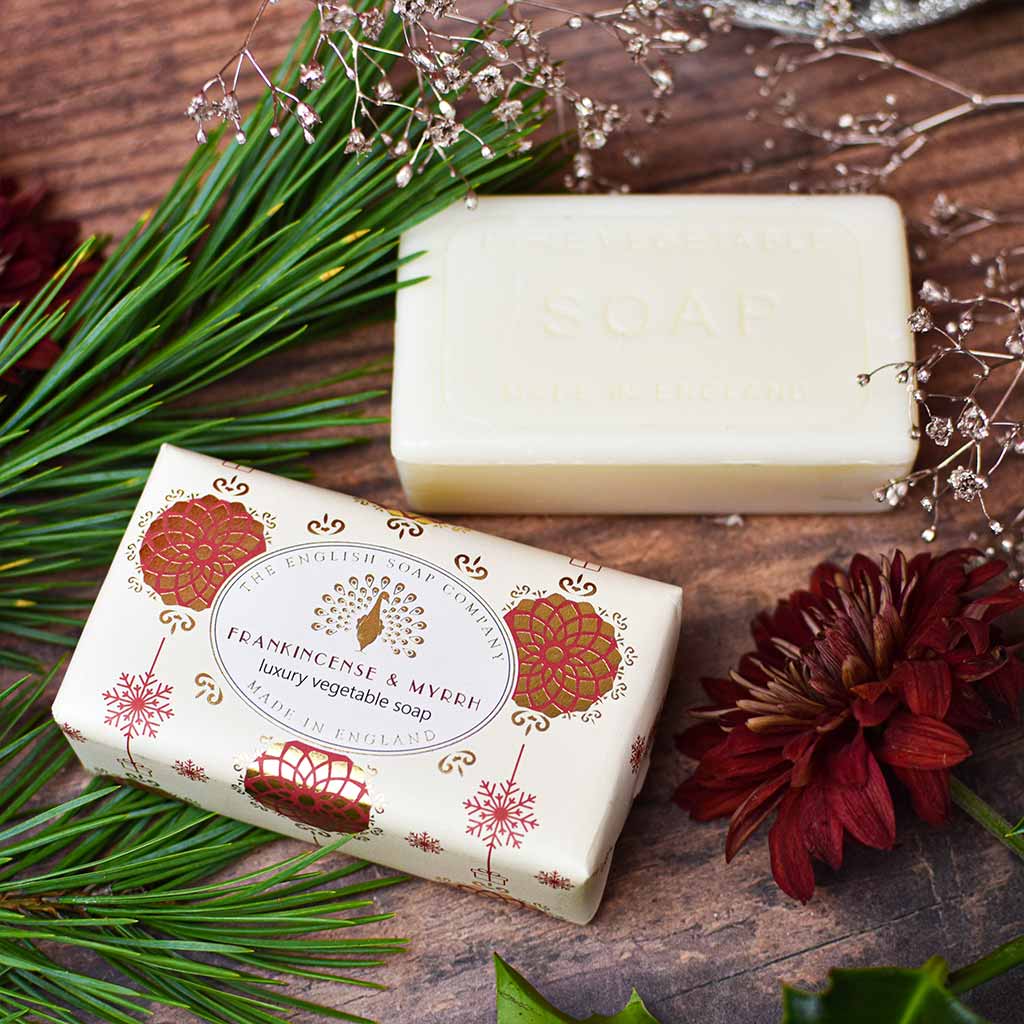 Two bars of English Soap Company soap arranged on a rustic wooden surface with green pine branches, dried flowers, and a dark red flower. One bar is wrapped in cream paper with ornate red and gold botanical illustrations and the words 'FRAMPINCET & MYERY luxury vegetable soap'; the other bar is unwrapped, showing a solid creamy off-white soap bar with 'SOAP' embossed on top.