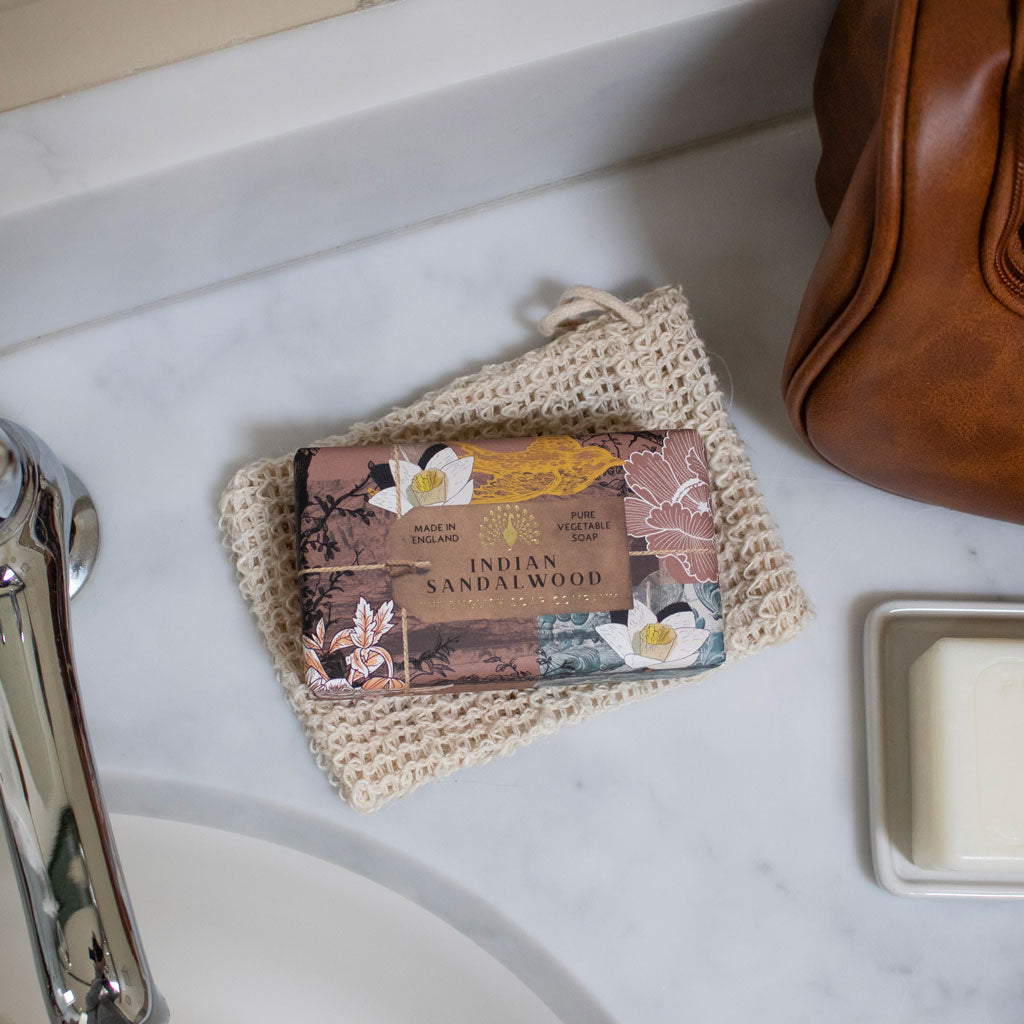 Bar of soap with 'Indian Sandalwood' label on a textured cloth next to a brown leather bag on a marble surface.