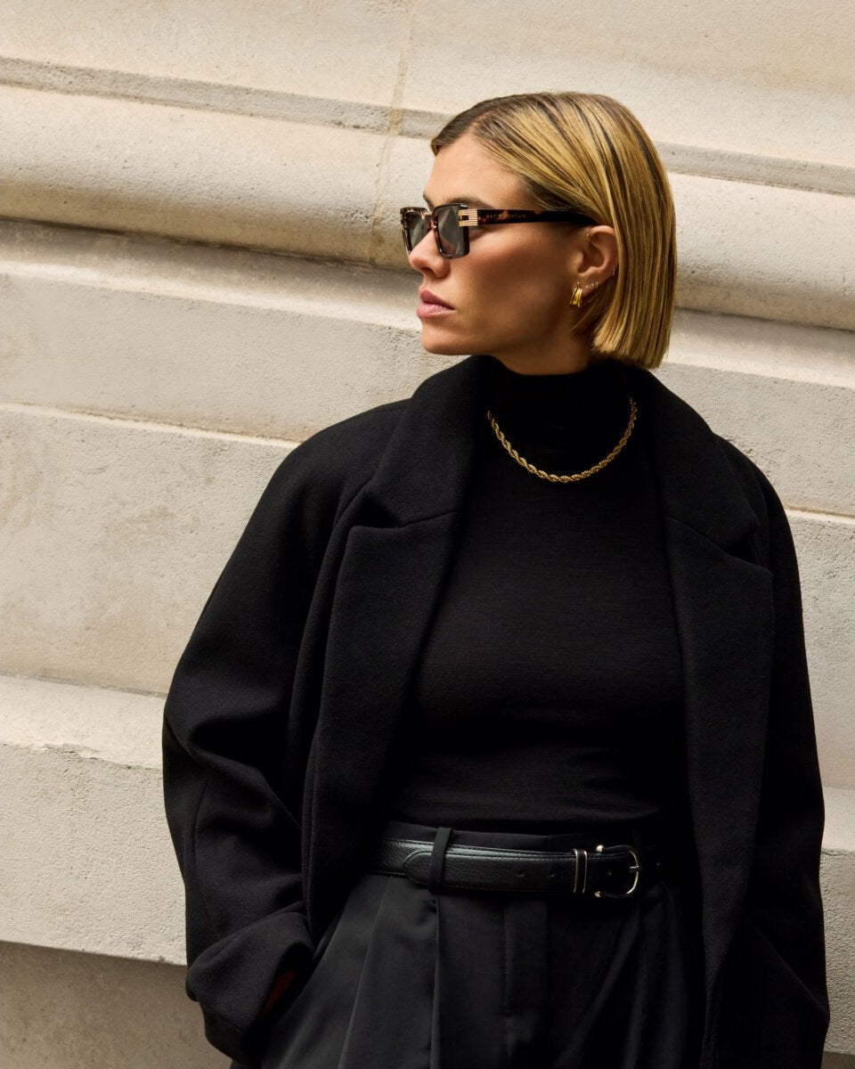 Woman in a black coat wearing katie loxton malibu tortoiseshell sunglasses standing against a light-colored wall
