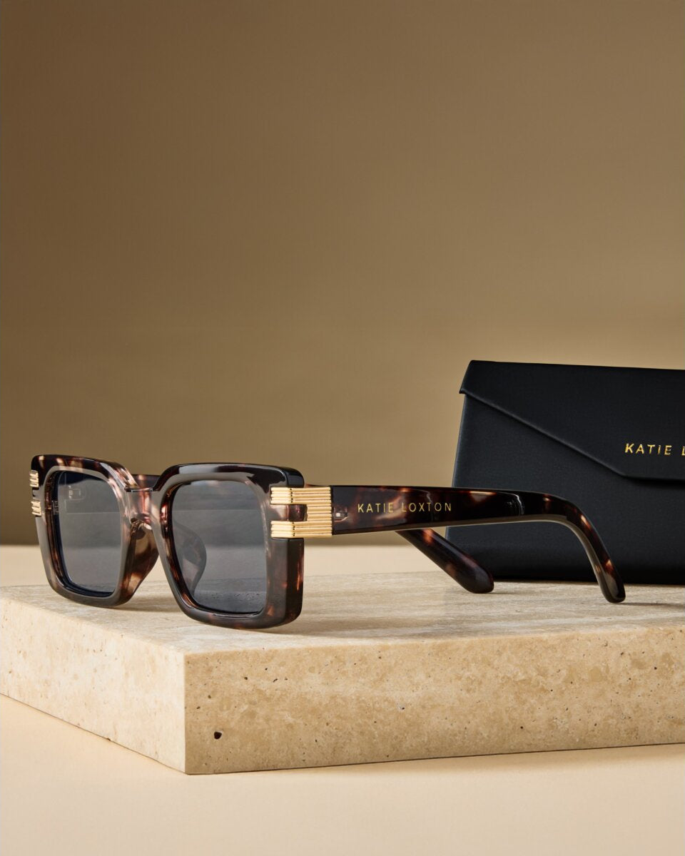 katie loxton malibu Sunglasses with tortoiseshell frame and gold accents on a beige surface, with a matching case in the background.