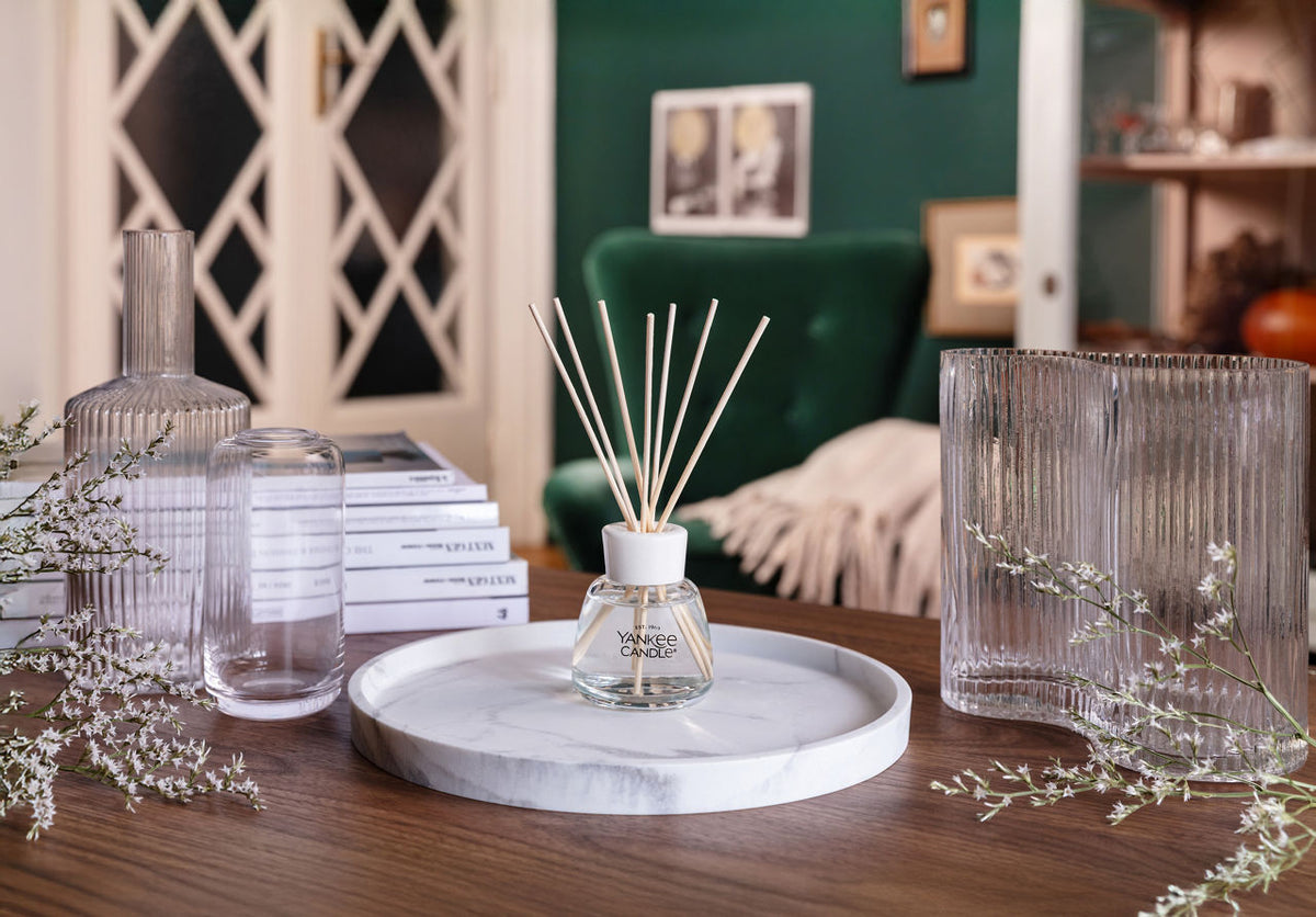 A clear glass reed diffuser with light wooden reeds sits on a round white marble tray on a wooden table, styled with glass bottles, books, and dried flowers in a cozy room setting.
