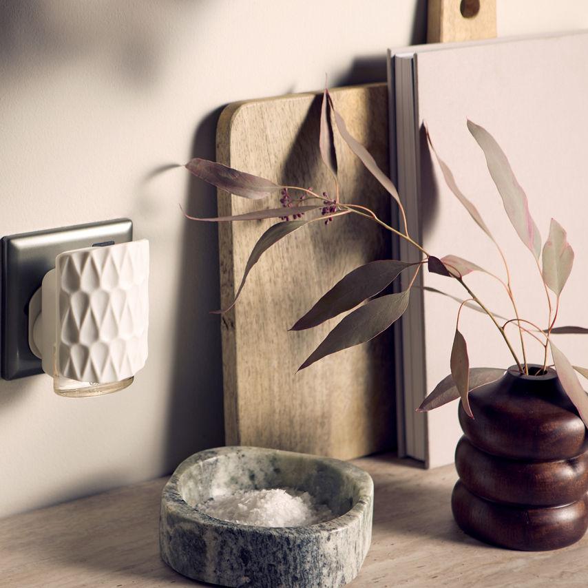scent plug inserted into wall socket, surrounded by glassware and a vase on a surface with a neutral background.