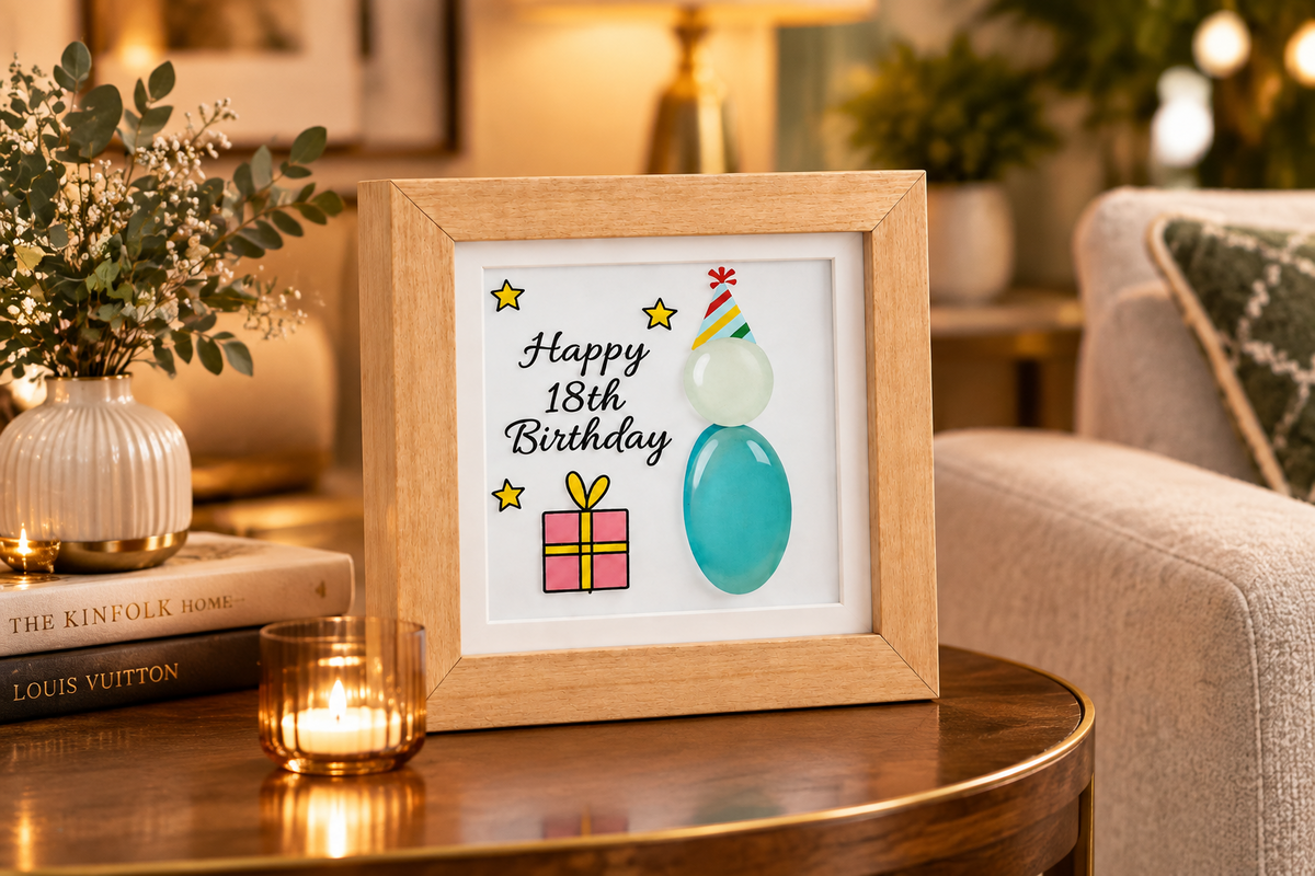 A square framed 18th birthday pebble frame  on a wooden table in a cozy living room setting. The print features a light wood frame with a white background, displaying the text 'Happy 18th Birthday'