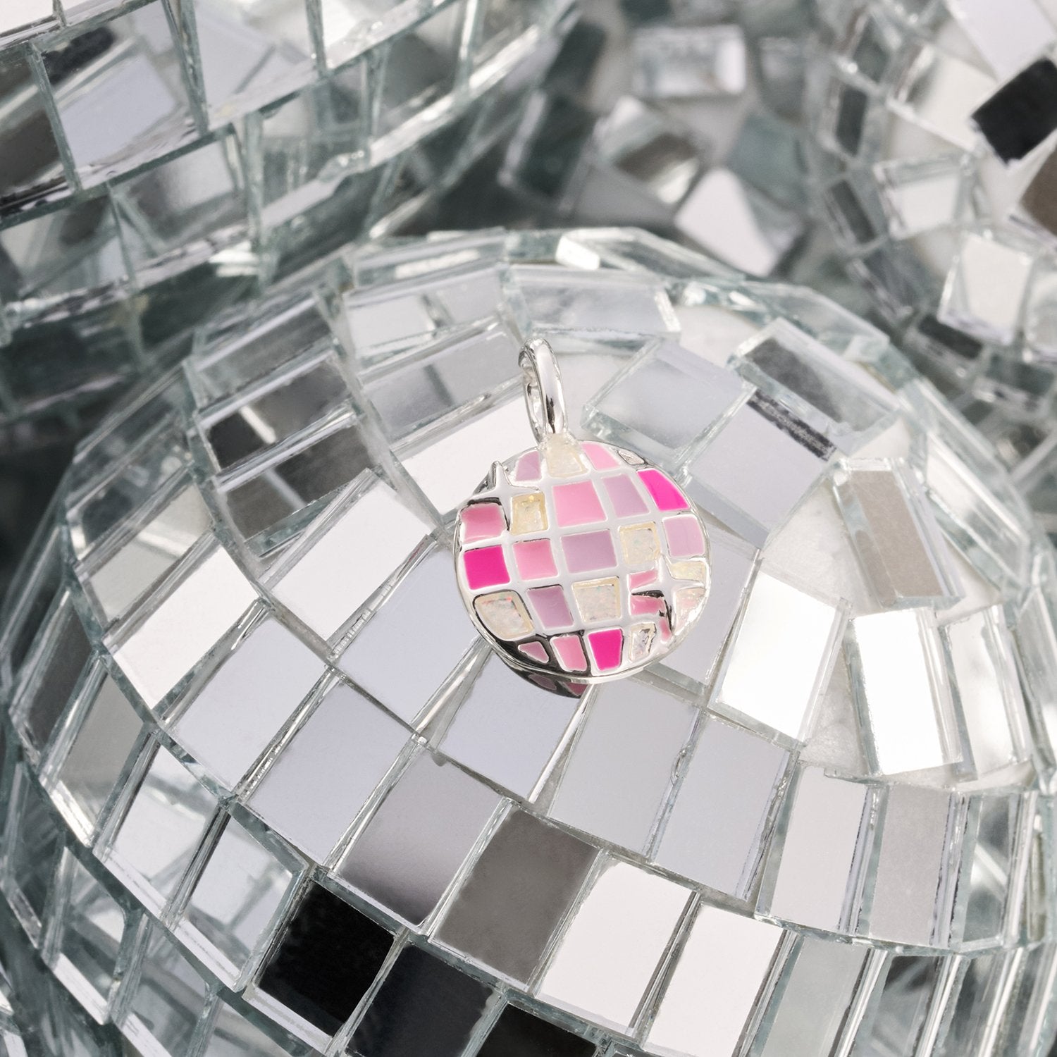 A small round disco ball pendant charm with pink, white, and silver mirrored tiles, resting on a larger mirrored disco ball surface.