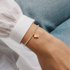 A close-up of a person's wrist wearing a delicate Joma Jewellery silver beaded bracelet with a small pink and gold heart charm on a chocolate bar