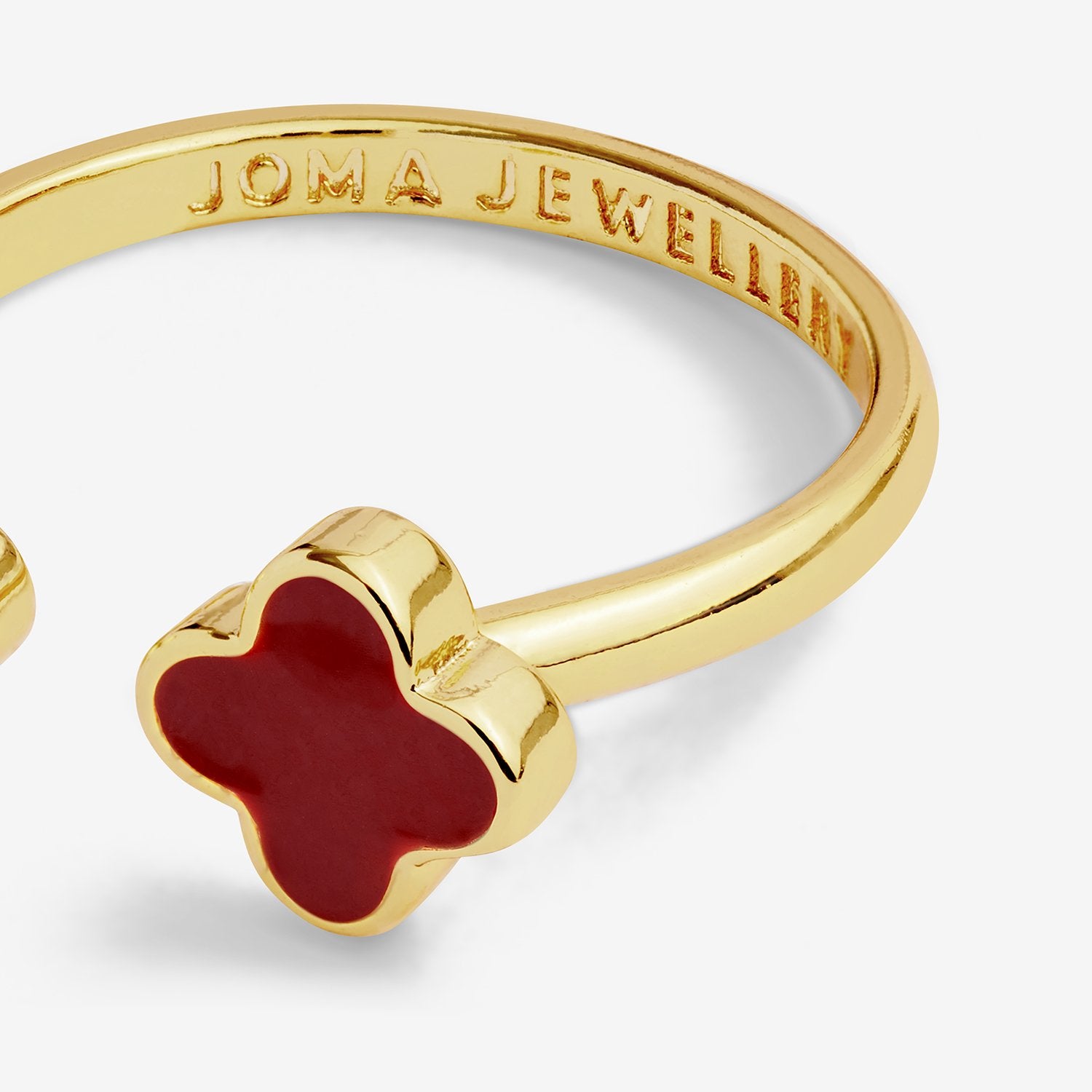 Gold-plated open cuff ring featuring a clover-shaped charm with a deep red enamel center, shown on a light gray background. The inside of the band is engraved with 'JOMA JEWELLERY'.
