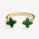 A gold open cuff ring by Joma Jewellery, featuring two four-pointed flower motifs in green malachite inlay, shown on a light gray background.
