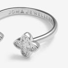 A silver-tone open bangle ring by Joma Jewellery, featuring a polished four-petal flower charm set with clear stones on the front, shown on a clean white background.