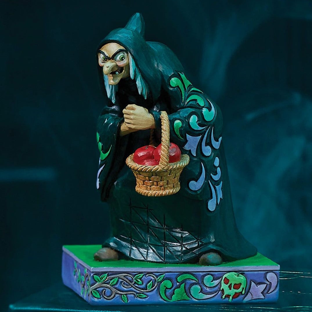 A decorative figurine of a witch in a dark teal robe holding a basket of red apples, standing on a patterned base against a dark blue background.