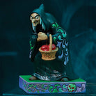 A decorative figurine of a witch in a dark teal robe holding a basket of red apples, standing on a patterned base against a dark blue background.