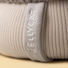 Close-up of a Jellycat amuseables pomilly hat with a gray cable-knit top, a cream ribbed bottom, a gray pom-pom, and small fabric hands and feet, shown on a white background with brand name 'JELLYCAT' embroidered vertically on a fabric tab.
