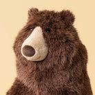 A close-up of jellycats Bizzlestomp brown plush bear toy with a cream-colored snout and dark eyes, set against a solid pale yellow background.