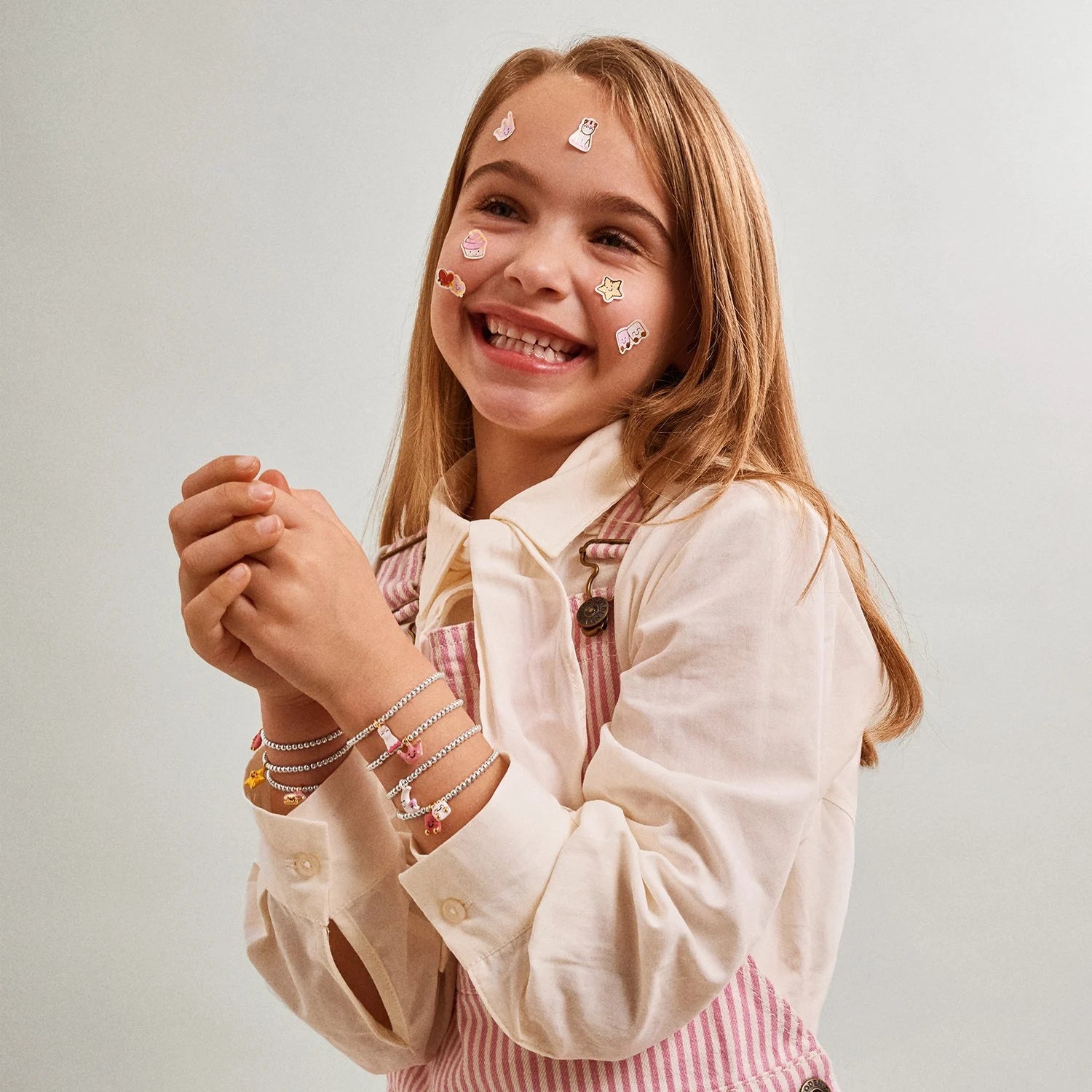 A young girl with heart-shaped face stickers smiles while wearing multiple delicate silver-plated Jomees's bracelets on her wrists.