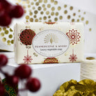 A rectangular bar of luxury vegetable soap from The English Soap Company, labeled 'Frankincense & Myrrh', wrapped in cream paper with red and gold foil patterns, displayed on a white block with festive props.