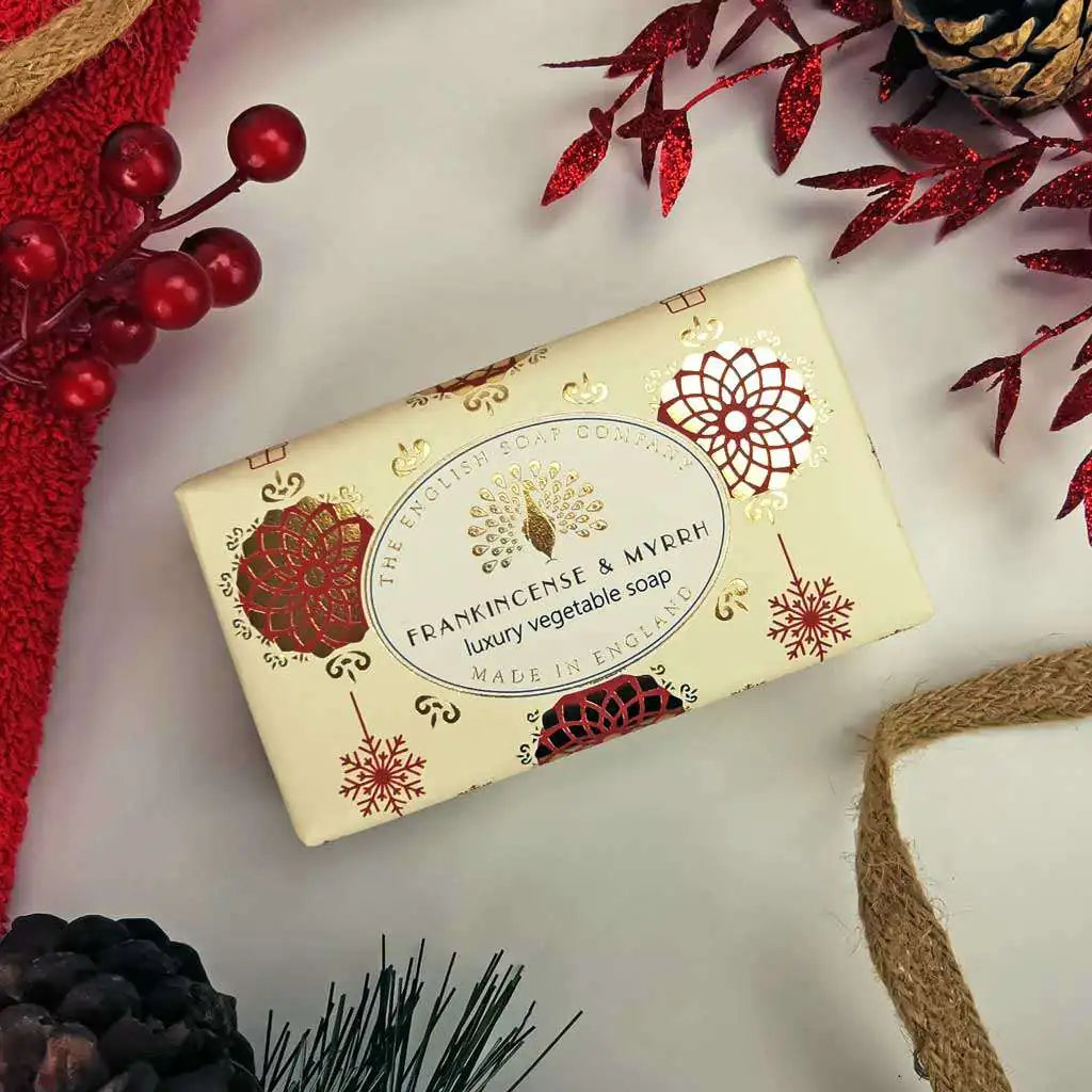 A rectangular bar of Frankincense & Myrrh soap by The English Soap Company, wrapped in cream-colored paper with red and gold festive illustrations, styled on a white surface with holiday decorations.