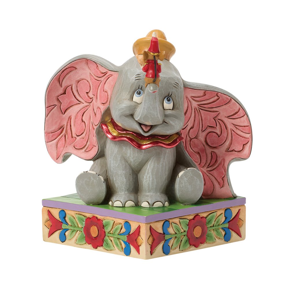 A decorative figurine of Dumbo the elephant sitting on a patterned base, with pink wings, a red and gold collar, and a small gold crown, photographed on a plain white background.