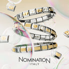 Three silver-toned Nomination Italy charm bracelets featuring  yellow gold links from the composable Nomination charm collection. Spiral on a soft cream background with floating bubbles and the text 'NOMINATION ITALY' at the bottom.