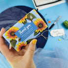Hand holding a soap bar with sunflower design and 'Thank you' text on a blue background