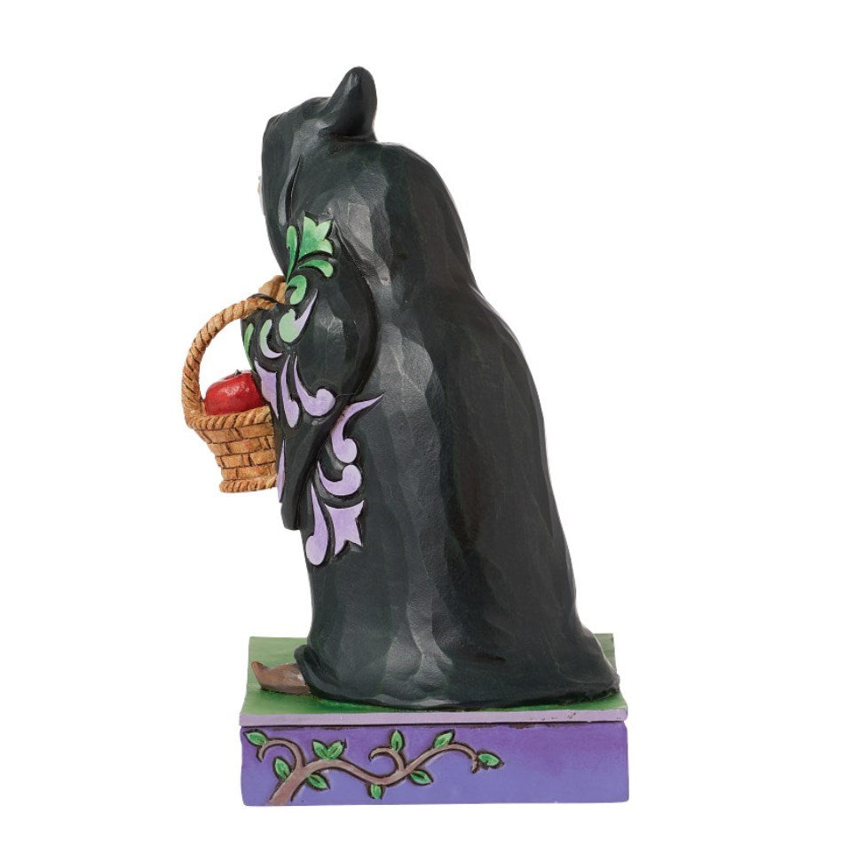 A decorative figurine of a witch seen from the back, holding a small woven basket with a red apples.  standing on a purple base decorated with a branch and leaves.