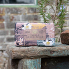 A rectangular bar of Indian Sandalwood pure vegetable soap with a floral and landscape patterned wrapper, resting on a rustic wooden crate in front of a blurred brick wall and greenery.