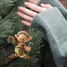 A hand wearing a ribbed gray sweater hangs a small Christmas ornament shaped like a gnome elf in a green pine tree. The ornament wears a red Santa hat and a beige outfit with red and green floral details, holding a candy cane and a blue ball.