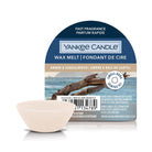 A Yankee Candle amber & sandalwood wax melt package with a small beige wax melt chip in front, on a clean white background. The package is dark blue with a beach scene and driftwood graphic.