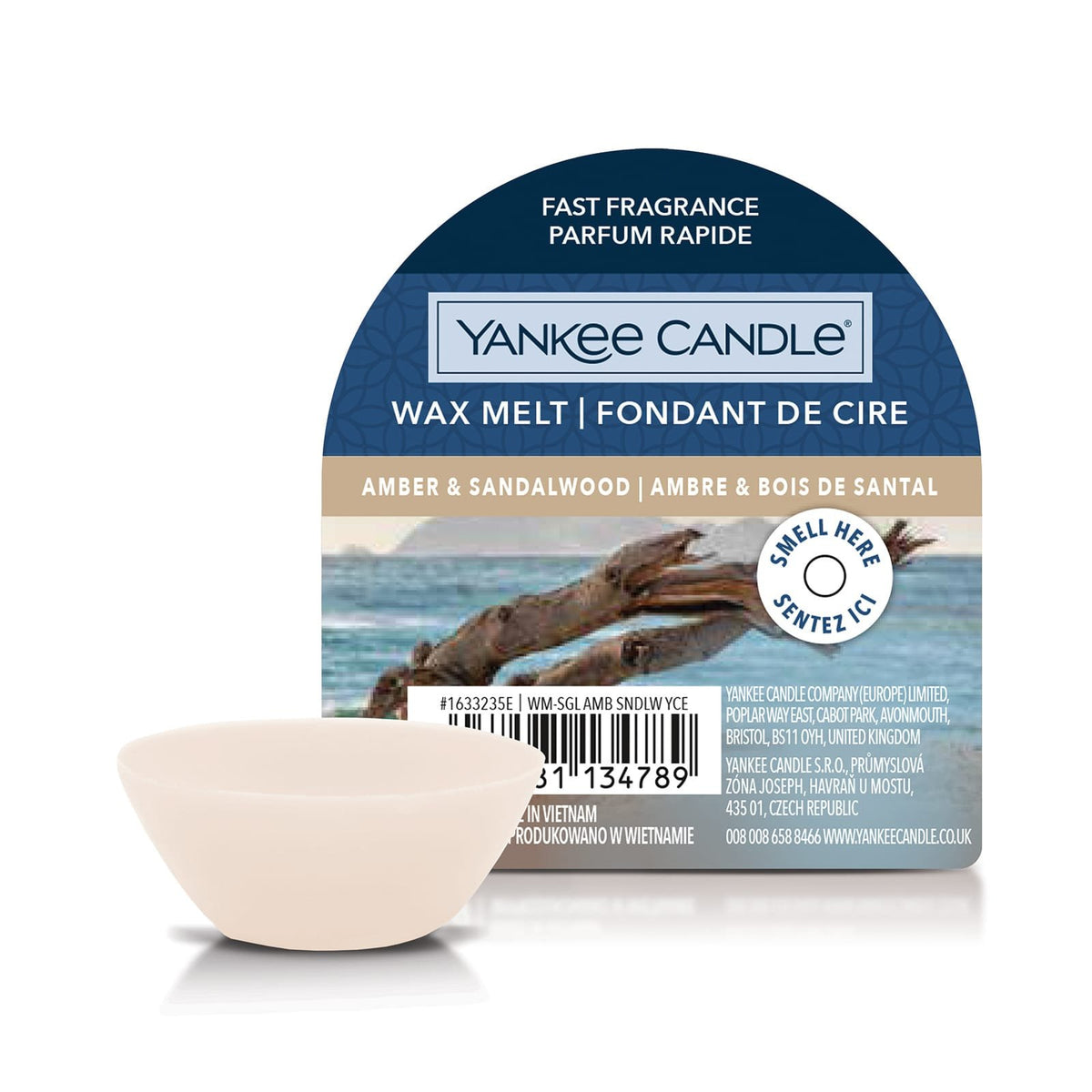 A Yankee Candle amber & sandalwood wax melt package with a small beige wax melt chip in front, on a clean white background. The package is dark blue with a beach scene and driftwood graphic.
