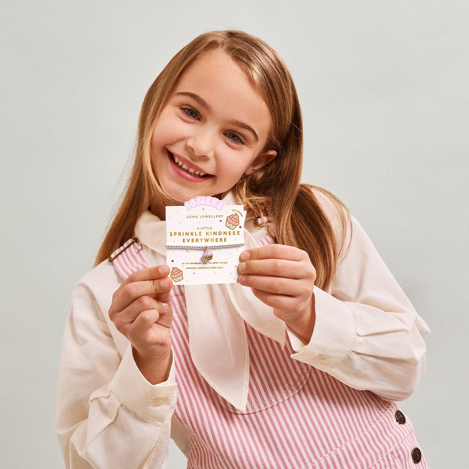 Young girl holding a sprinkle kindness everywhere childrens jomee bracelet. On its presentation gift card with text against a plain background