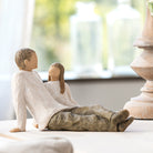 displayed Willow tree figurine, of a father and his daughter sitting together. On top of a a table with a blurred background scene 