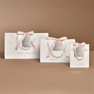Three white Joma Jewellery gift bags in different sizes, each tied with a pale pink satin ribbon and bow, arranged on a warm beige background.