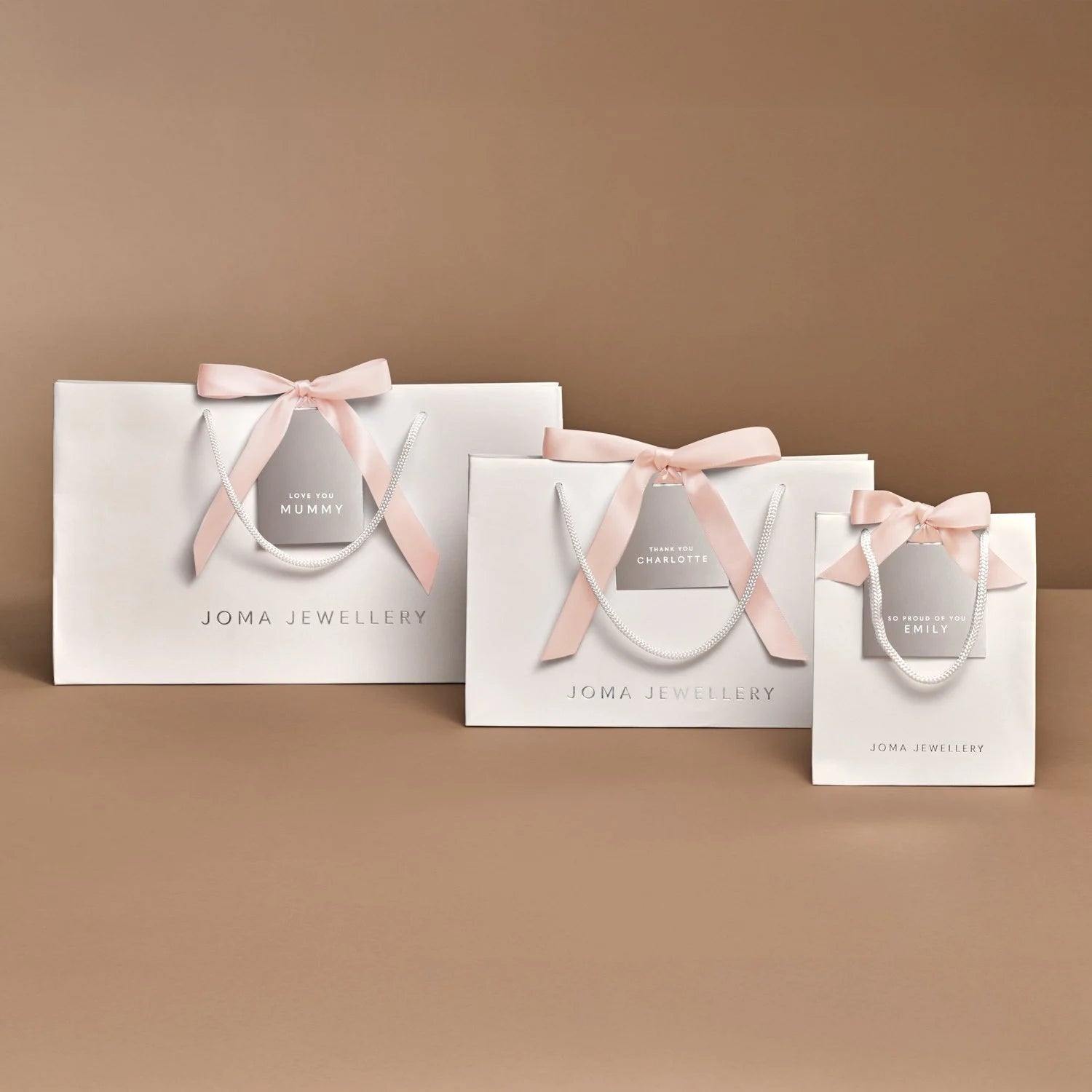 Three white Joma Jewellery gift bags in different sizes, each tied with a pale pink satin ribbon and bow, arranged on a warm beige background.