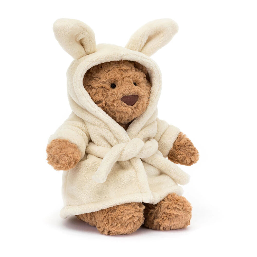 A plush jellycat Bartholomew teddy bear wearing a cream-colored hooded bathrobe, photographed against a clean white background.