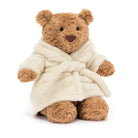 A brown plush jellycat Bartholomew teddy bear wearing a cream-colored plush bathrobe, photographed against a solid white background.