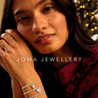 A close-up lifestyle shot of a woman wearing three delicate silver bracelets from Joma Jewellery littles collection. These include a silver plated heart-shaped charm with pave, a small heart another with a pave circle. She is wearing a dark red ruffled blouse, with a warm, softly blurred holiday background.