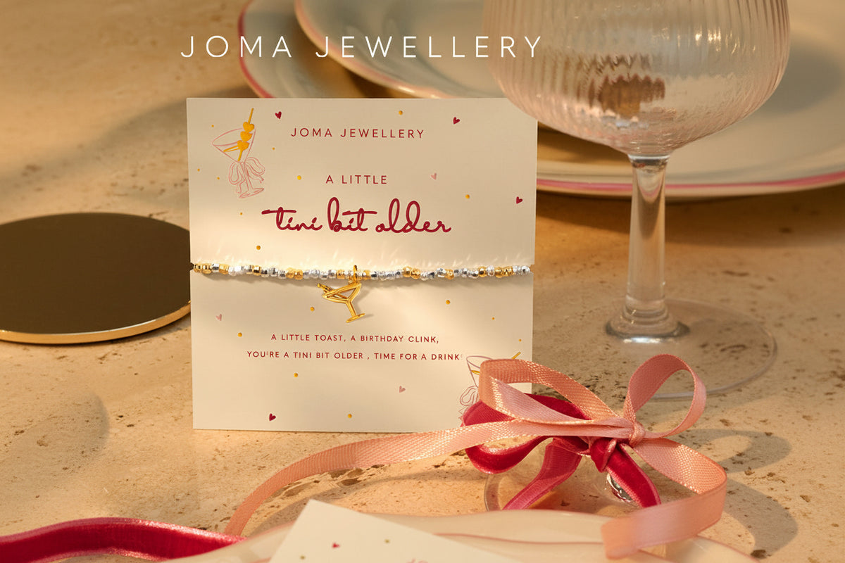 A Joma Jewellery bracelet with a gold-toned charm and silver glitter band, presented on a branded cream card with pink and gold accents, styled on a tabletop with ribbons and glassware.