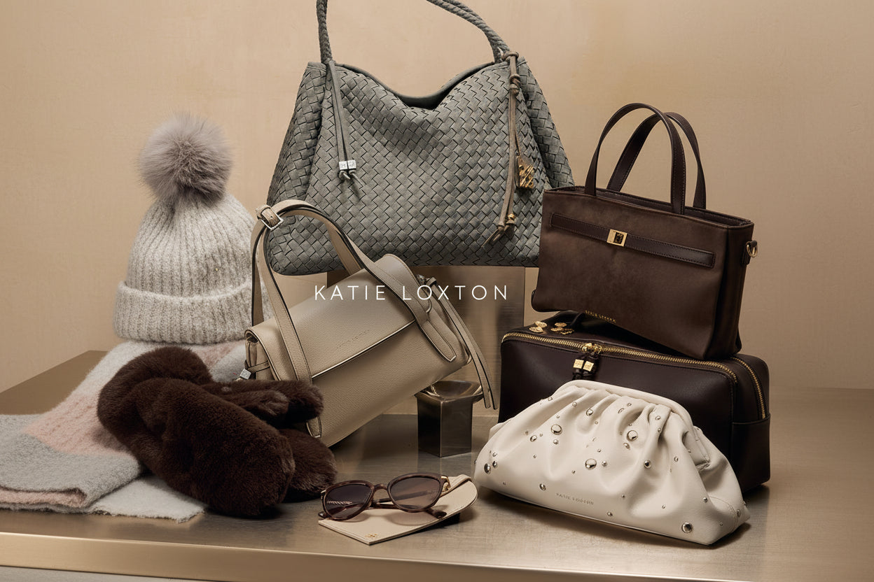 A styled collection of Katie Loxton handbags arranged on a tabletop, including a large woven gray shoulder bag, a light beige leather bag, a dark brown bag, a cream pouch with metal studs, sunglasses, and a knit hat, with the text 'KATIE LOXTON' overlaid in the center.