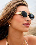 Woman wearing gold katie loxton siena matte sunglasses with a blurred beach background