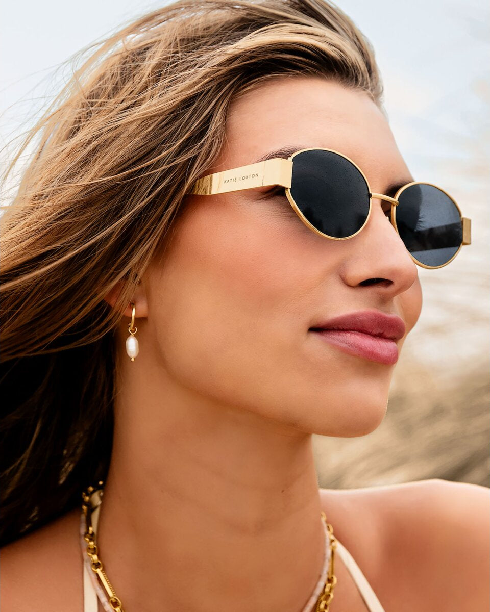 Woman wearing gold katie loxton siena matte sunglasses with a blurred beach background