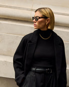 Woman in a black coat wearing katie loxton malibu tortoiseshell sunglasses standing against a light-colored wall