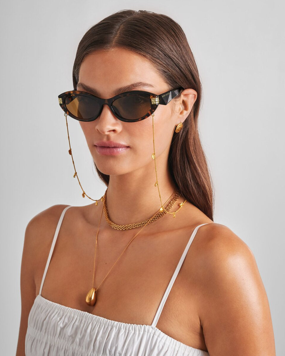 Woman wearing katie loxton sunglasses with a chain, gold necklace, and white top on a gray background