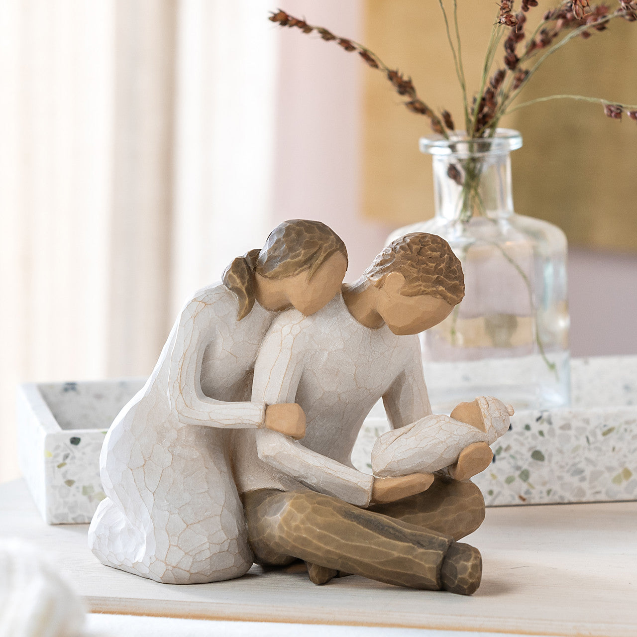 willow tree Decorative figurine, of a woman sitting  leaning into a male. While he's holding a child on a table with a vase in the background