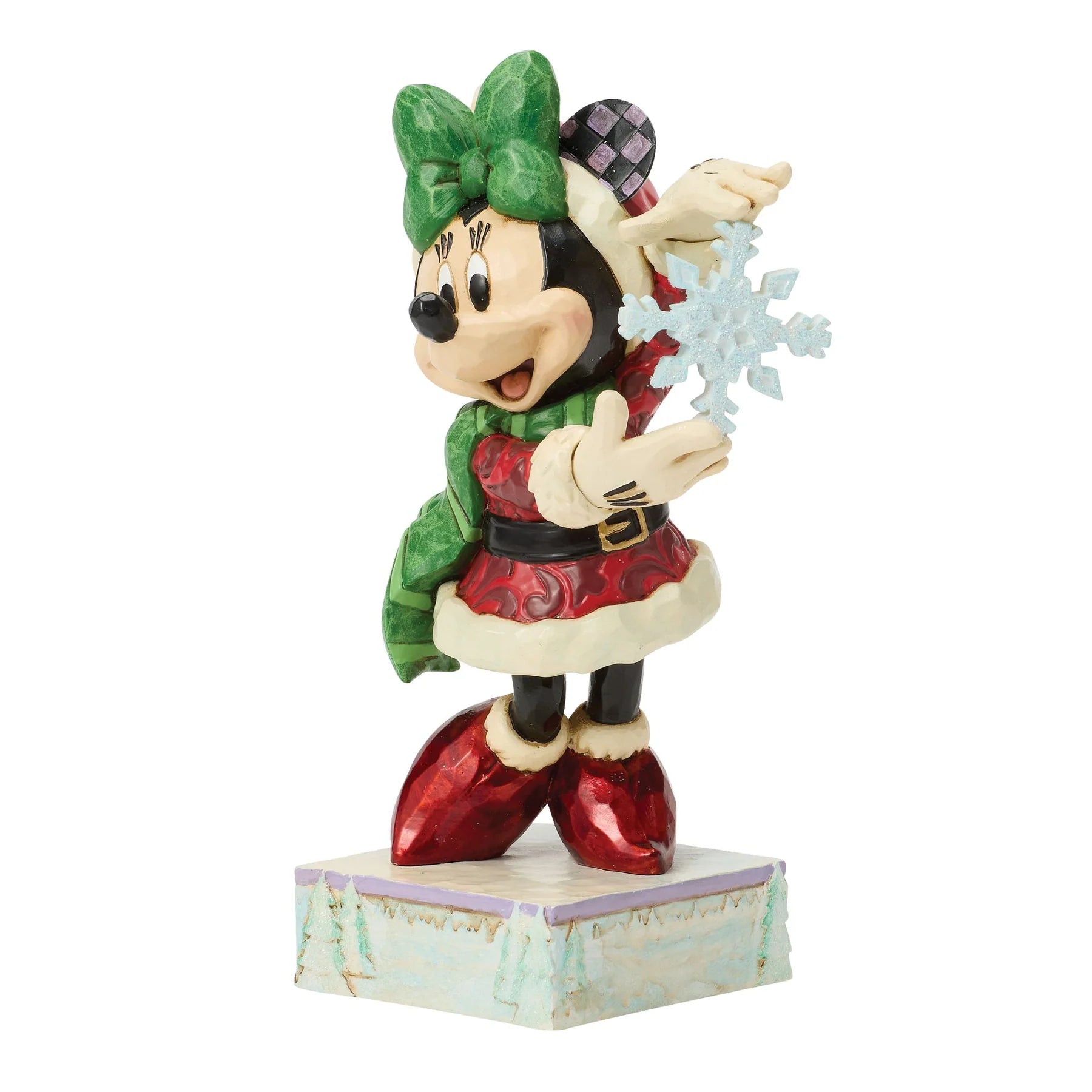 A Disney Minnie Mouse holiday figurine wearing a green bow, red and green Christmas outfit, and red boots, holding a light blue snowflake while standing on a decorative base against a white background.
