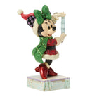 A Disney Minnie Mouse holiday figurine wearing a green bow and scarf, red dress, and red boots, holding a silver ornament on a decorative base against a white background.