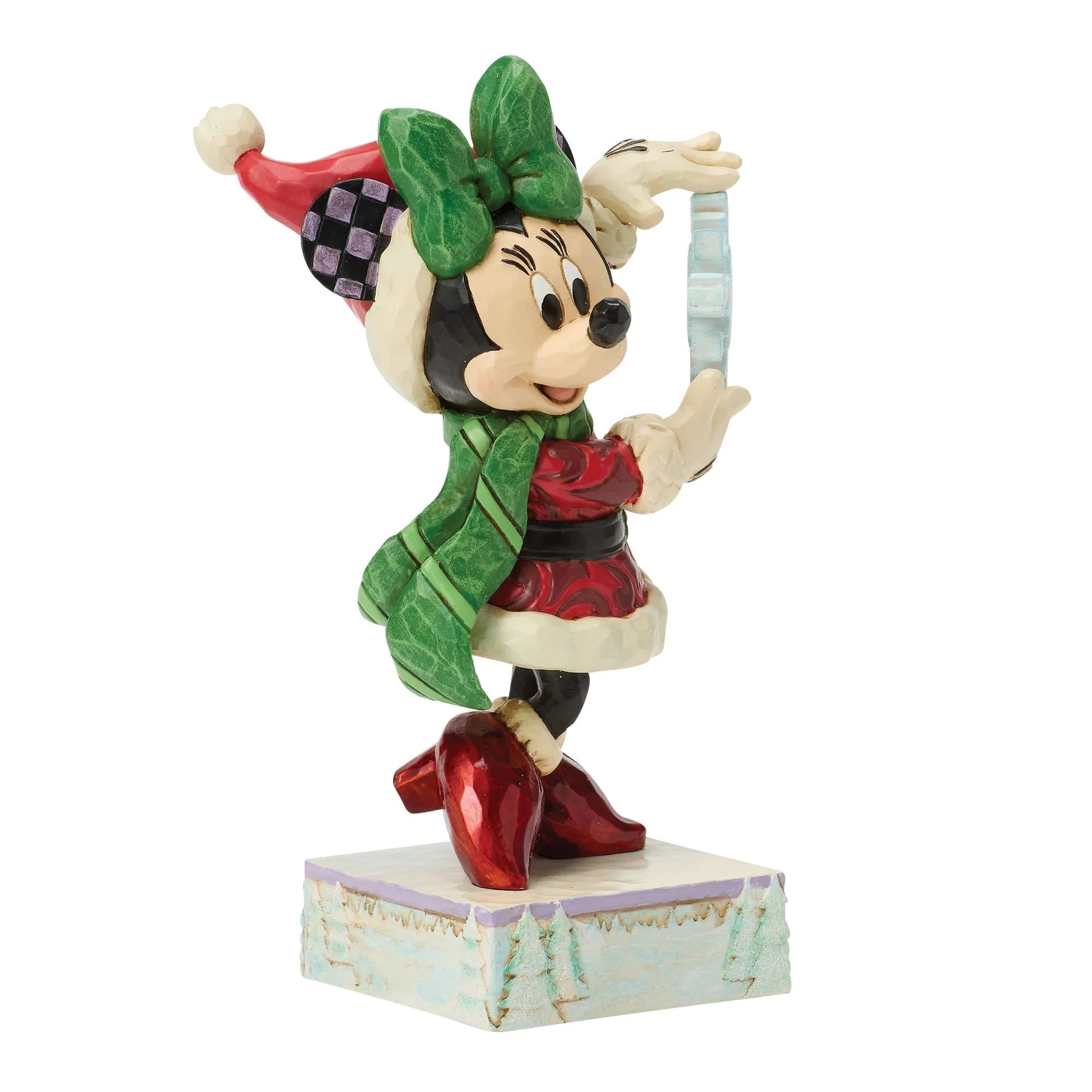 A Disney Minnie Mouse holiday figurine wearing a green bow and scarf, red dress, and red boots, holding a silver ornament on a decorative base against a white background.