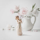 willow tree beautiful wishes figurine holding a vase with flowers in front of a larger vase with pink flowers on a light back ground